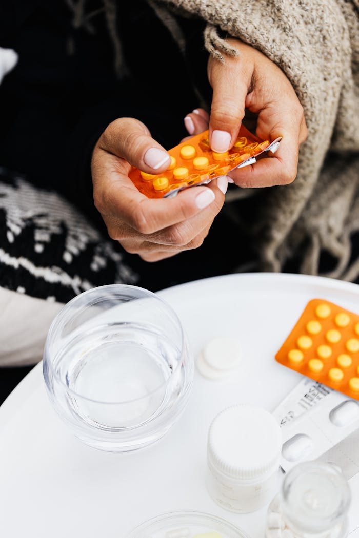 Close-up of hands handling blister pack with yellow pills. Includes glass of water on table.