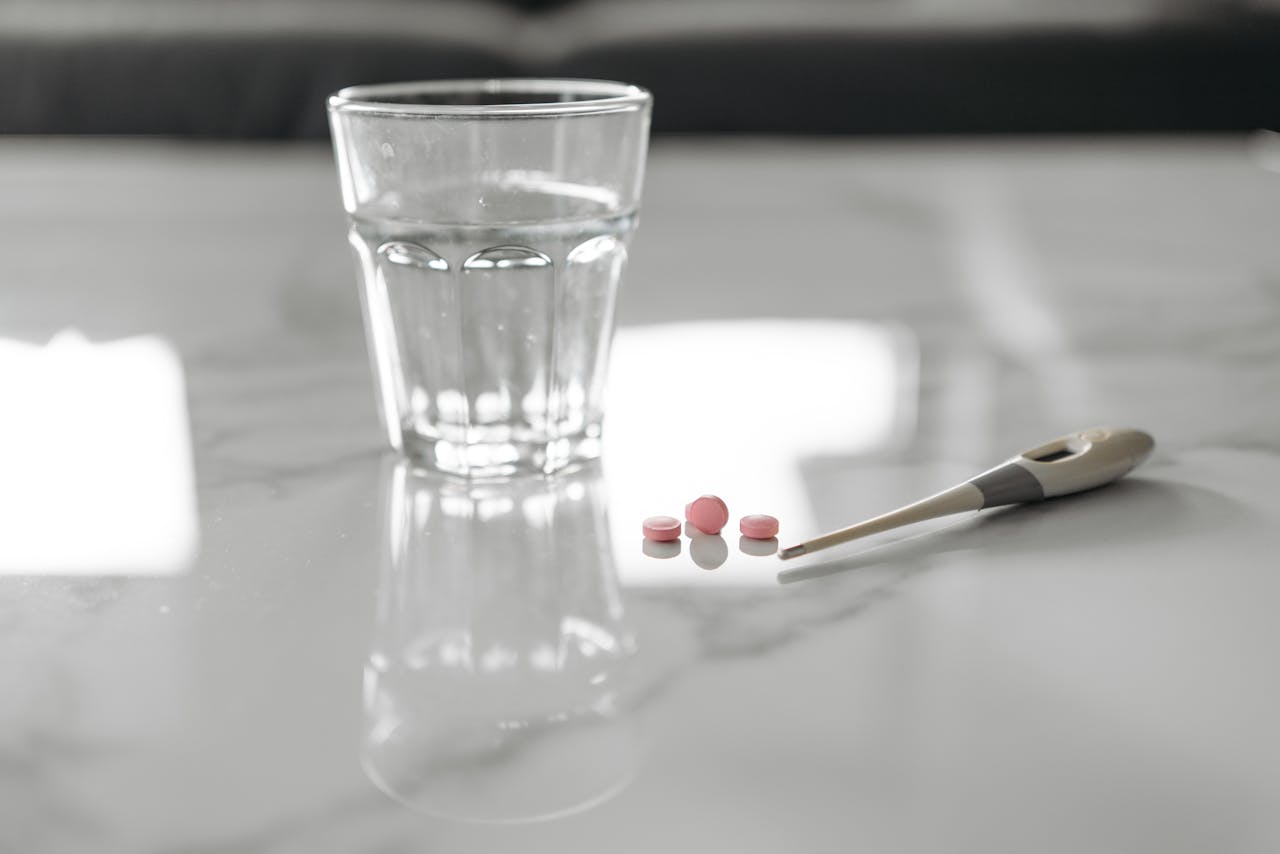 A glass of water and pills next to a thermometer on a reflective marble surface.